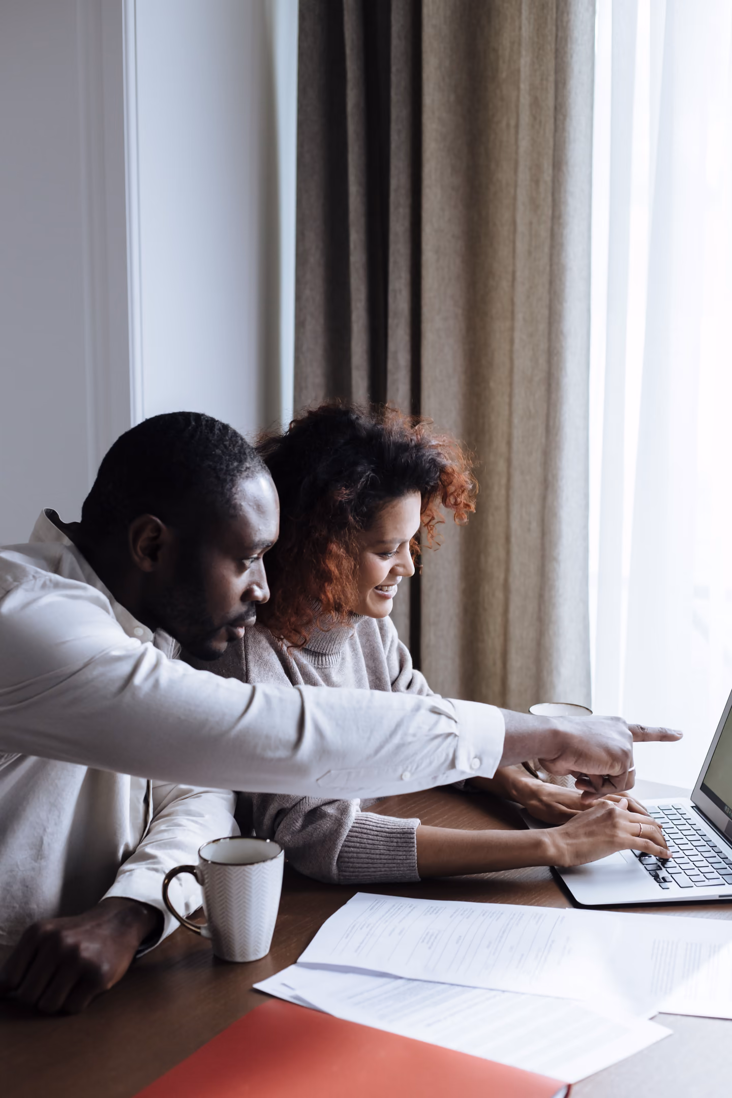 A couple at a kitchen table, looking at a laptop together, papers spread between them.