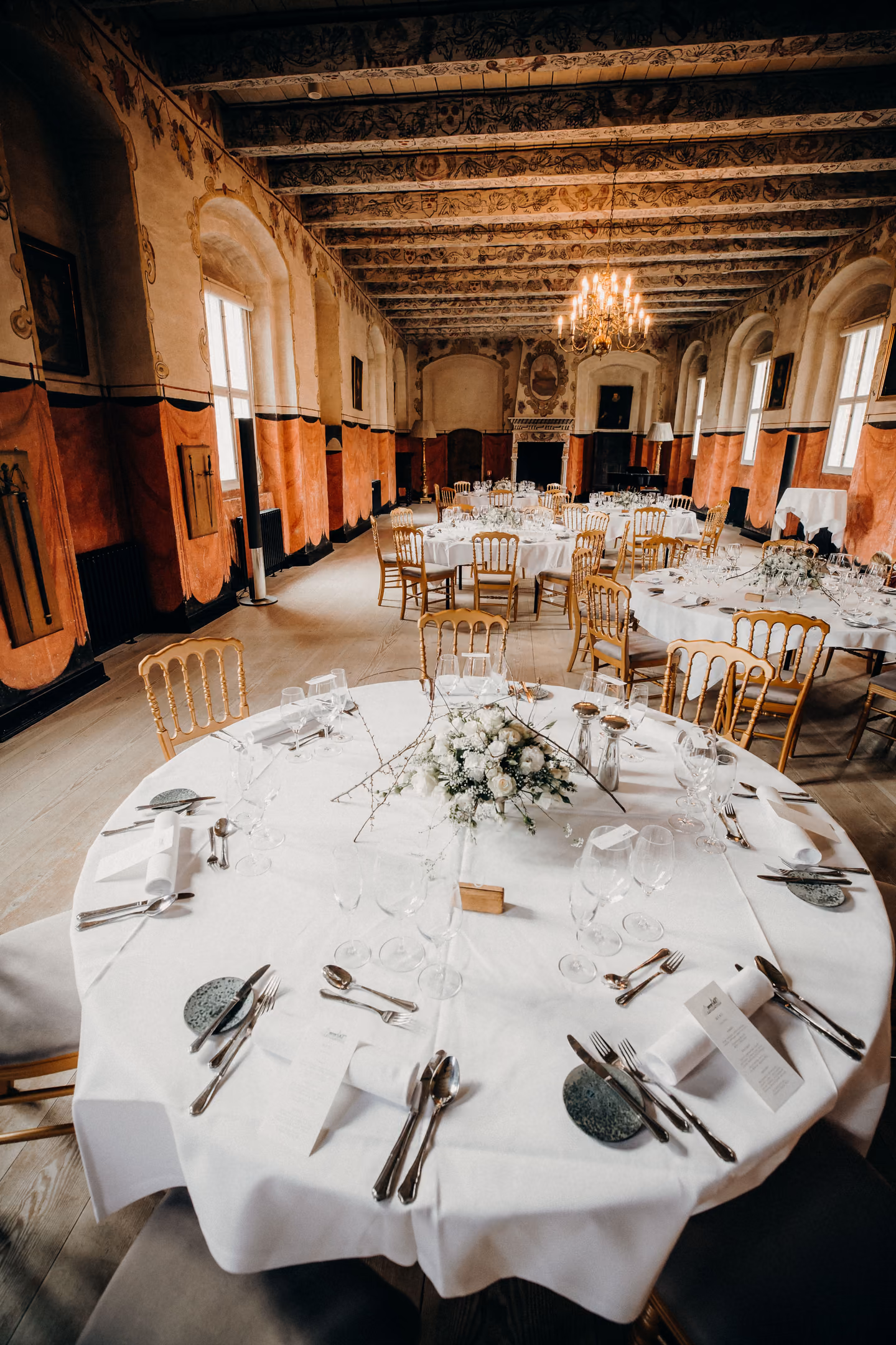 Round tables set with glasses and plates in an ornate hall with tall windows and a painted ceiling.