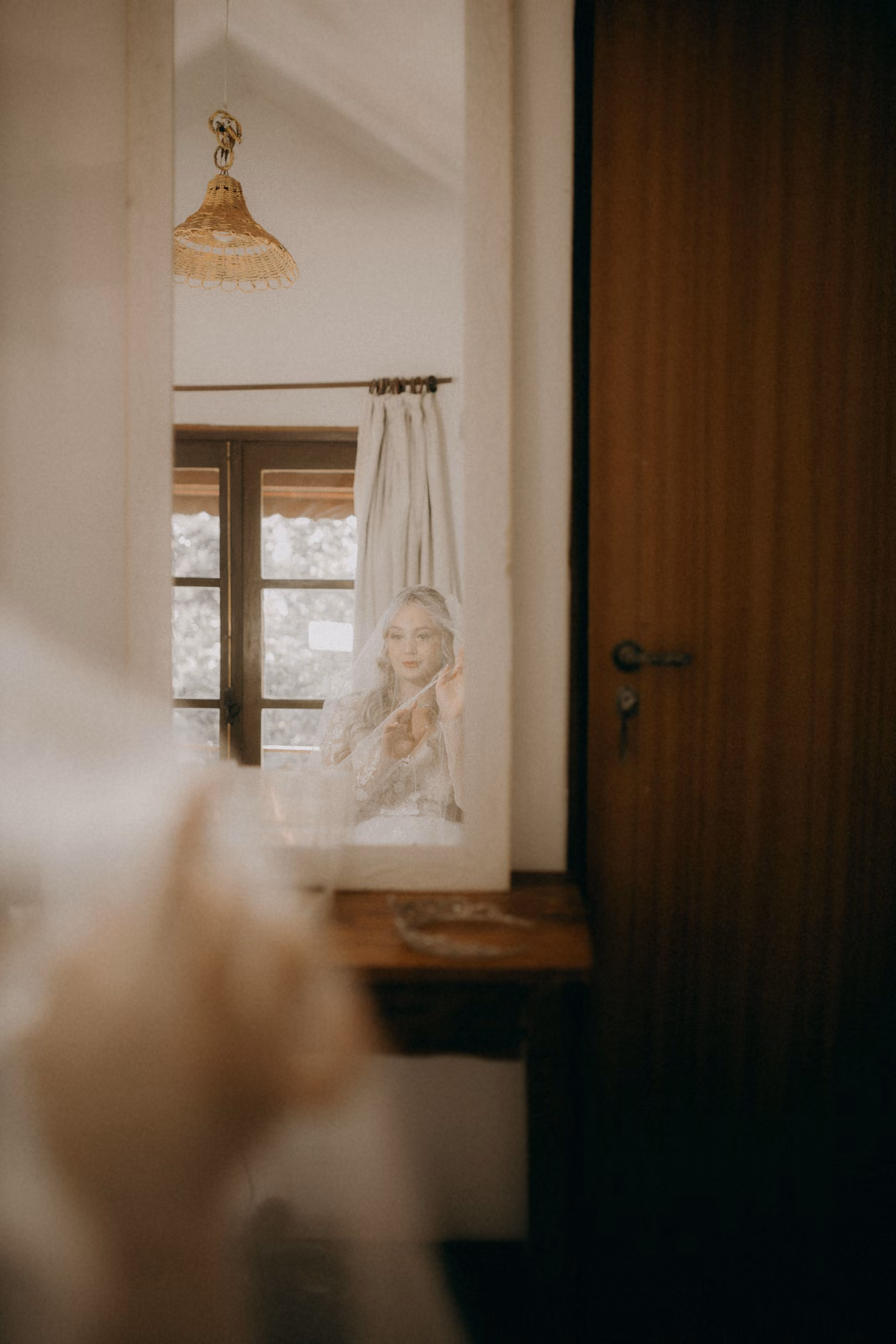 A bride seen in a mirror while getting ready in a quiet room, morning light from a window.