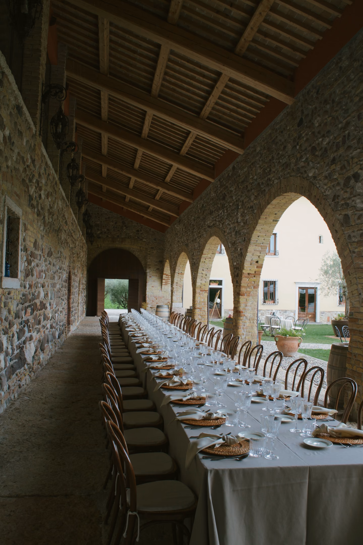 A long dinner table set for many guests under stone arches, empty chairs along both sides.